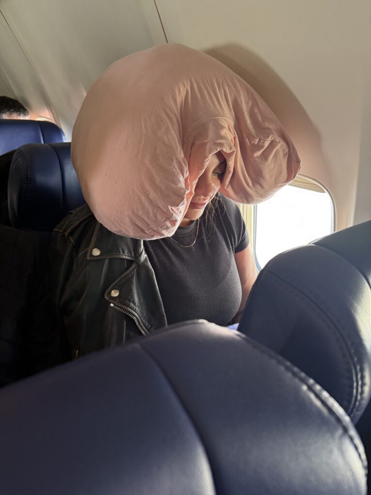 A woman, sitting on a plane, with a Sleep Crown on her head and leather jacket slung over her shoulders